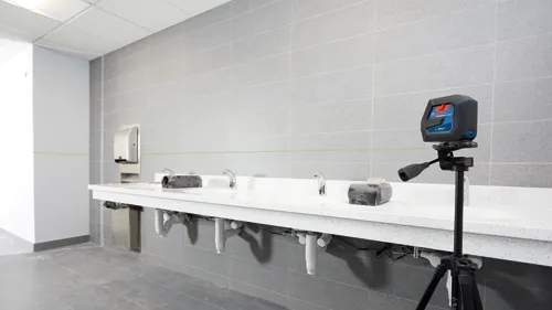 Niveau laser Bosch Professional GCL 100-40G sur trépied, projetant des lignes laser vertes dans des toilettes publiques.