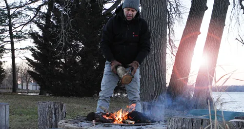 Une personne portant un sweat à capuche noir avec le mot "Bosch" sur la poitrine, un pantalon gris et un bonnet gris tient une bûche au-dessus d'un feu.
