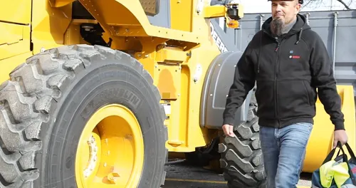Homme portant une veste noire avec le logo "BOSCH", marchant près d'un engin de chantier jaune avec un pneu "MICHELIN".