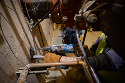 Perceuse à percussion rotative Bosch Bulldog Xtreme Flex, avec un opérateur portant des gants perçant du béton près de tuyaux.