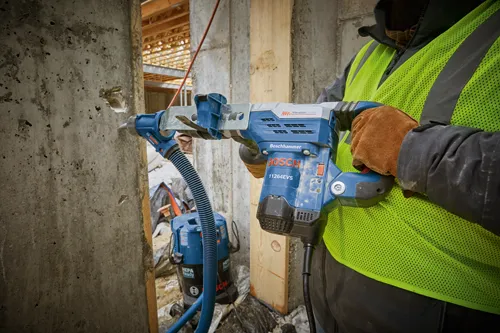 Bosch 11264EVS Boschhammer avec VibrationControl, utilisé par une personne portant un gilet de sécurité et des gants. L'outil est connecté à un aspirateur avec le texte "HEPA Ready".