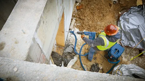 Voici la traduction :

Une personne portant un gilet réfléchissant et un casque de chantier utilise un perforateur Bosch sur un chantier de construction, avec un aspirateur à proximité.