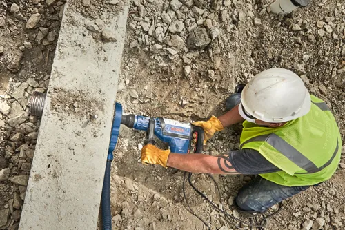 Voici la traduction :

Personne portant un casque de sécurité et un gilet de sécurité utilisant un Boschhammer RH745 Bosch pour travailler sur du béton.