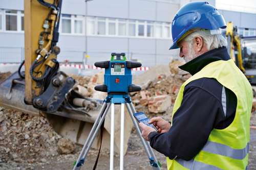 Homme utilisant un smartphone près d'un niveau laser rotatif Bosch sur un trépied, portant un casque bleu et un gilet de sécurité jaune, avec un arrière-plan de chantier de construction.