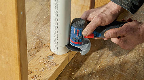 Meuleuse angulaire Bosch GWS12V-30 coupant un tuyau en PVC près d'un cadre en bois.