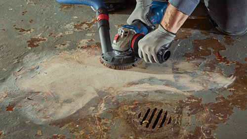 Une personne portant des gants utilise une meuleuse d'angle Bosch sur un sol, avec un tuyau d'aspirateur connecté à la meuleuse.