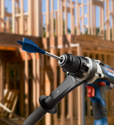 Perceuse Bosch avec une mèche plate bleue devant une ossature de bâtiment.