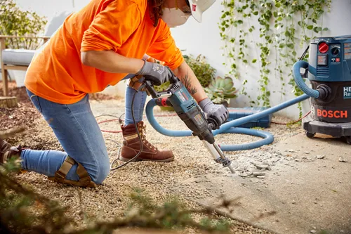 Personne agenouillée, portant une chemise orange à manches longues, un jean, des bottes de travail marron et des gants, utilisant un marteau perforateur Bosch Bulldog Xtreme connecté à un aspirateur de poussière HEPA Bosch.
