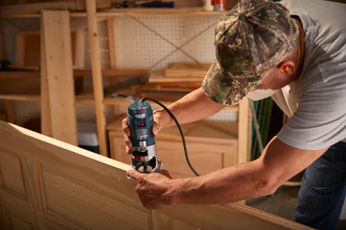 Routeur Bosch Colt 1.0 HP utilisé par une personne portant un chapeau de camouflage, des lunettes de sécurité et un masque facial.