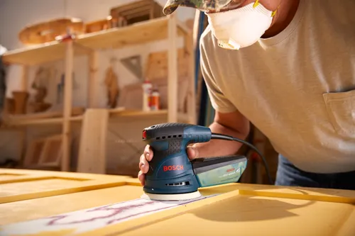 Voici la traduction :

Personne portant un masque utilisant une ponceuse Bosch, avec "BOSCH" et "système microfiltre" visibles sur la ponceuse.