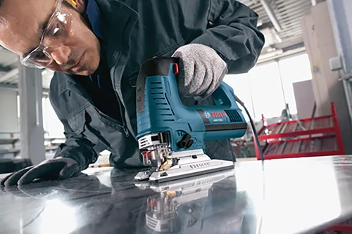 Homme portant des lunettes et des gants de sécurité utilise une scie sauteuse, avec la marque "BOSCH" dessus.