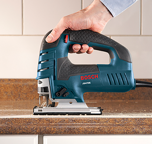 Scie sauteuse BOSCH JS470E, tenue par une personne à la peau claire, coupant un plan de travail marron.