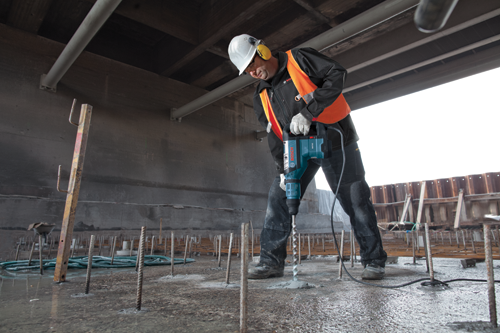 Homme portant un casque de protection, une protection auditive et un gilet de sécurité orange utilisant un marteau rotatif Bosch RH745 pour percer une surface en béton.
