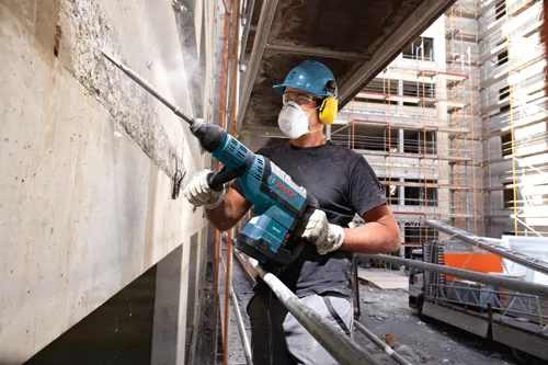 Une personne portant un équipement de sécurité utilise un marteau rotatif Bosch RH745 pour casser du béton, avec un chantier de construction en arrière-plan.
