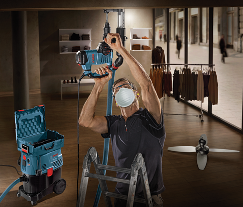 Homme utilisant un marteau perforateur rotatif Bosch, portant des lunettes de sécurité et un masque, debout sur une échelle, connecté à un aspirateur Bosch. Des étagères avec des vêtements et un portant avec des vêtements sont visibles en arrière-plan.