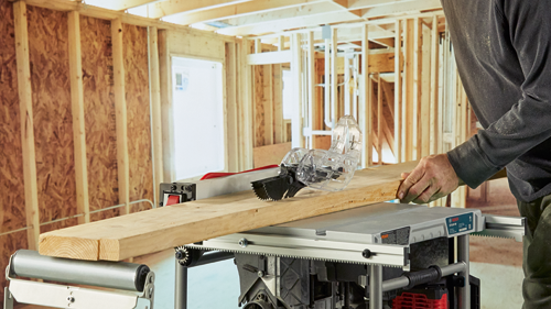 Scie à table Bosch GTS1041A-09, avec protection de sécurité, coupant une planche de bois ; une personne tient la planche ; arrière-plan de chantier.