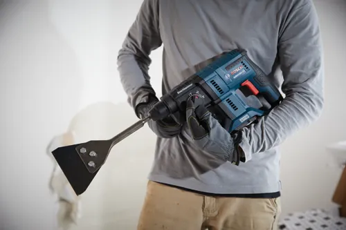 Bosch GBH18V-21 marteau perforateur, avec une mèche à burin attachée, tenu par une personne portant une chemise à manches longues et des gants.