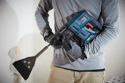 Perceuse à percussion rotative Bosch GBH18V-21, avec un accessoire burin, tenue par une personne portant des gants de travail et une chemise grise à manches longues.