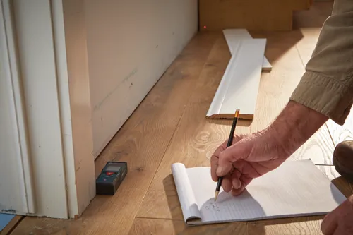 Une personne écrit sur un bloc-notes avec un crayon à côté de plinthes et d'un télémètre, "BOSCH" visible.