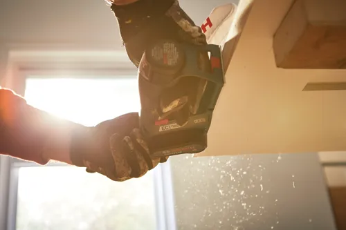 Bosch GHO12V-08 moteur sans balais, tenu par des mains gantées, coupant du bois ; poussière en suspension.