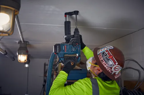 Voici la traduction :

Une personne portant des lunettes de sécurité et un casque de protection utilise un marteau rotatif Bosch RH328VC sur un plafond. La personne porte un équipement de sécurité et des gants RIDGE CUT.