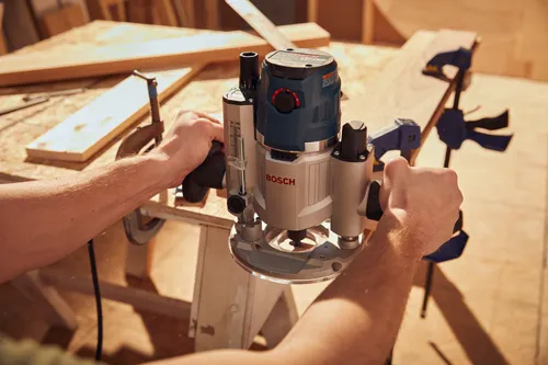 Voici la traduction :

Une personne utilise une défonceuse Bosch sur une surface en bois, fixée avec des serre-joints ; la défonceuse porte l'inscription « BOSCH » bien visible.