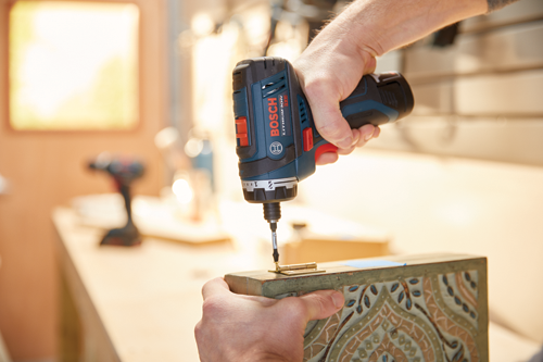 Une personne utilise une perceuse sans fil Bosch LITHIUM-ION 12V pour installer une charnière sur une boîte en bois.