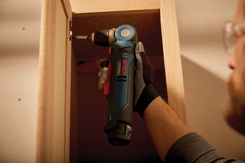 Une personne portant des lunettes de sécurité et des gants de travail utilise une perceuse Bosch Lithium-Ion 12V pour installer une porte de placard.