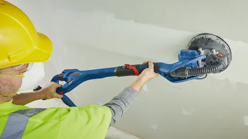 Une personne portant des lunettes de sécurité, un casque de chantier jaune et un gilet de sécurité vert vif, utilise une ponceuse à cloison sèche Bosch sur un mur blanc. La ponceuse est bleue et noire avec des touches de rouge.