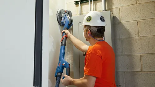 Voici la traduction :

Une personne portant des lunettes de sécurité, des bouchons d'oreille et un casque de protection utilise une ponceuse électrique Bosch pour poncer un mur. La personne porte un t-shirt orange.