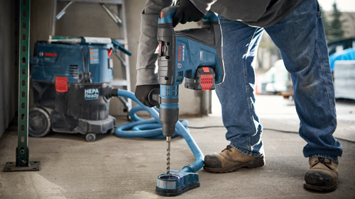 Personne portant des gants et un jean utilisant un perforateur Bosch GBH18V-40C avec un tuyau d'aspirateur raccordé.