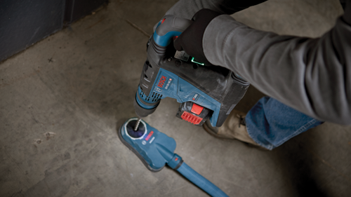 Personne portant une chemise grise à manches longues et des gants noirs tenant un marteau perforateur Bosch GBH18V-40C bleu et noir connecté à un aspirateur de poussière.