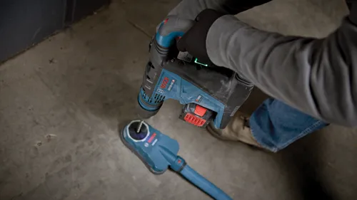 Personne portant une chemise grise à manches longues et des gants noirs tenant un marteau perforateur Bosch GBH18V-40C bleu et noir connecté à un aspirateur de poussière.