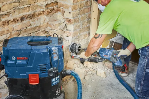 Une personne portant un masque et des gants utilise un perforateur, connecté à un aspirateur Bosch VAC090A HEPA Ready, pour percer un mur de briques.