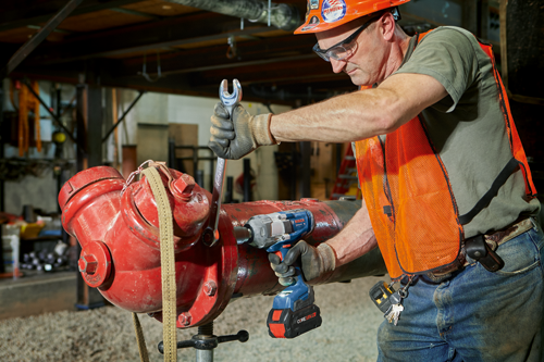 Une personne portant des lunettes de sécurité, un casque de protection et un gilet de sécurité orange, utilisant une clé et une visseuse à chocs Bosch GDX18V-1800 C Professional pour travailler sur une borne d'incendie rouge.