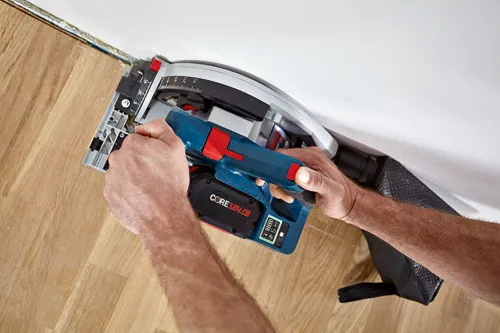 Scie circulaire sans fil Bosch, avec batterie CORE18V 8Ah, tenue par une personne coupant un plancher en bois près d'un mur.