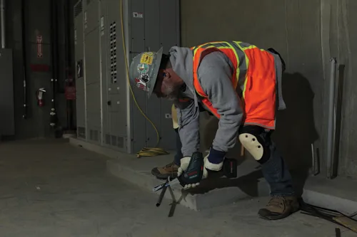 Une personne portant un casque de sécurité gris avec des autocollants, un gilet de sécurité orange et des genouillères coupe un tuyau métallique avec une scie alternative Bosch GSA18V-083.
