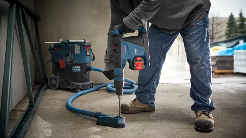 Voici la traduction :

Personne tenant un marteau perforateur rotatif Bosch GBH18V-40C, perçant une surface avec un aspirateur Bosch HEPA Ready en arrière-plan.