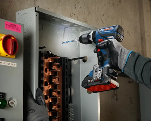 Bosch GSB18V-535C perceuse sans fil perçant dans une boîte électrique métallique, main gantée tenant une batterie CORE 18V 4Ah.