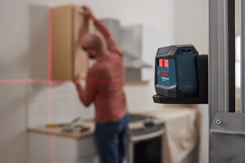 Bosch Professional GLL50-20, un outil de nivellement, projetant des lignes laser rouges horizontales et verticales ; une personne installe un meuble.