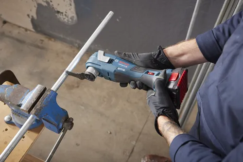 Bosch GOP18V-34 HD, outil multifonction oscillant à moteur sans balais, coupant un tuyau métallique fixé dans un étau ; une personne porte des gants noirs et une chemise bleu marine.