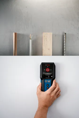 Détecteur Bosch Professional tenu par une main sur un mur blanc, au-dessus d'un mur gris avec un tuyau en cuivre, un câble, du bois et une barre d'armature.