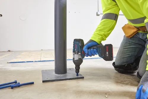 Bosch GDS18V-1000C clé à chocs, batterie CORE18V 2.0Ah, personne en équipement de sécurité utilisant la clé sur un poteau métallique, deux boulons bleus au premier plan.