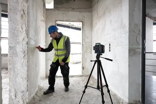 Voici la traduction :

Une personne portant un casque bleu, des lunettes de sécurité et un gilet réfléchissant utilise un niveau laser Bosch pour vérifier un mur de construction.