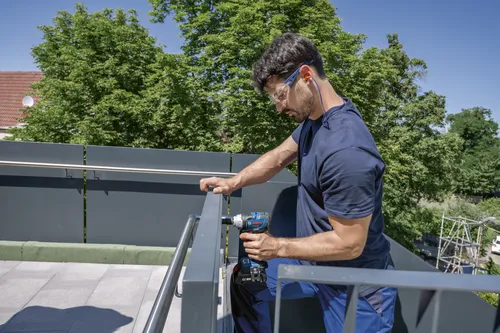 Homme utilisant une visseuse à percussion Bosch GSB18V-23C sur une rampe métallique, portant des lunettes de sécurité et une protection auditive.