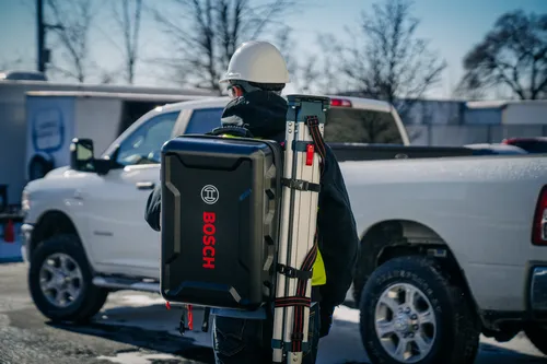 Personne portant un casque de sécurité blanc et un gilet de sécurité, transportant une mallette Bosch noire et un trépied, debout près d'un camion blanc.