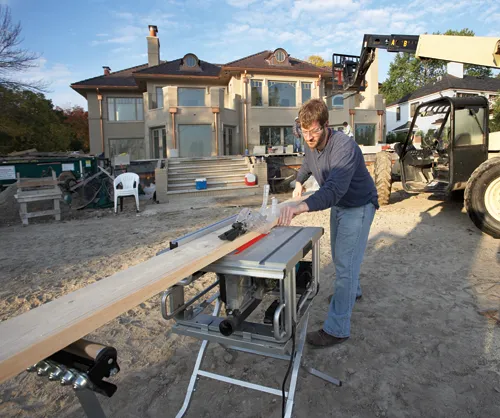 Homme portant des lunettes et une chemise bleue coupant du bois avec une scie à l'extérieur ; une grande maison et un véhicule de chantier sont en arrière-plan.