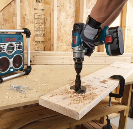Perceuse sans fil Bosch utilisant une scie cloche dans du bois, avec une radio Bosch sur le comptoir.
