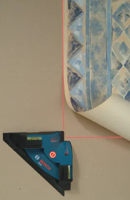 Niveau laser Bosch GTL2 projetant une ligne rouge sur un mur, près d'un rouleau de papier peint à motifs.