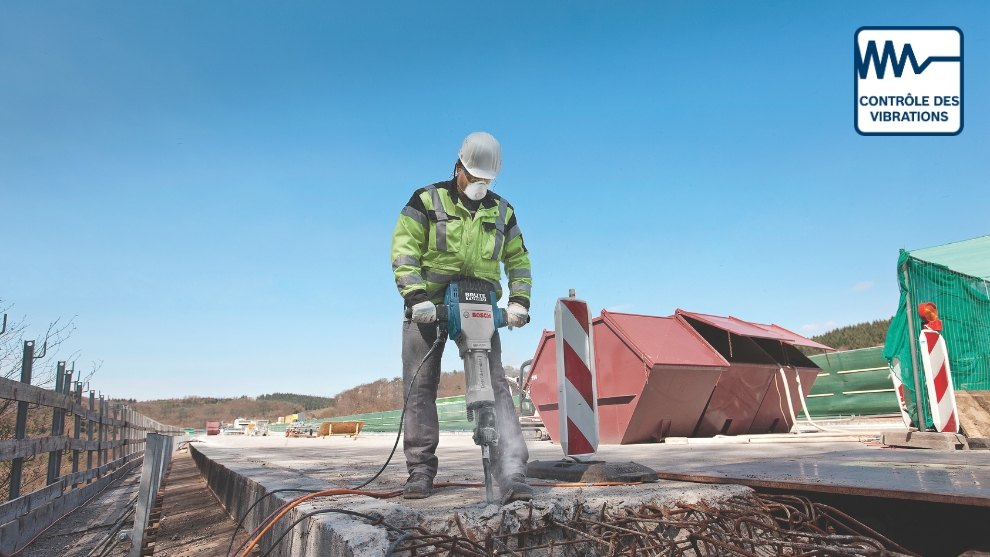 Un ouvrier du bâtiment en veste verte et noire utilise un marteau-piqueur Bosch Professional sur du béton, entouré de matériaux de construction et de machines sous un ciel bleu clair.